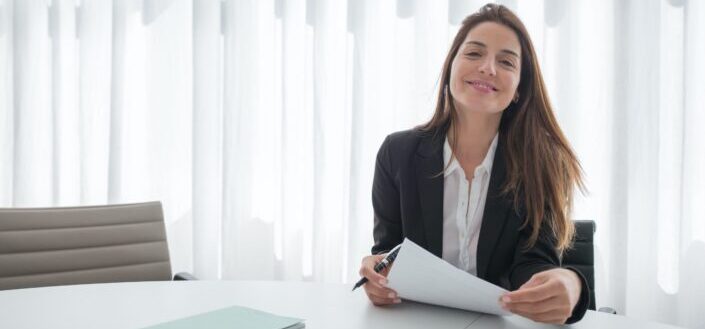 woman smiling in black blazer holding white paper and pen