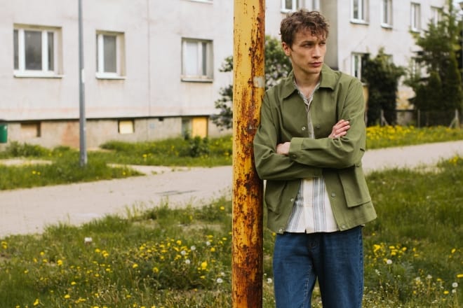 omega male - man in jacket leaning on a post