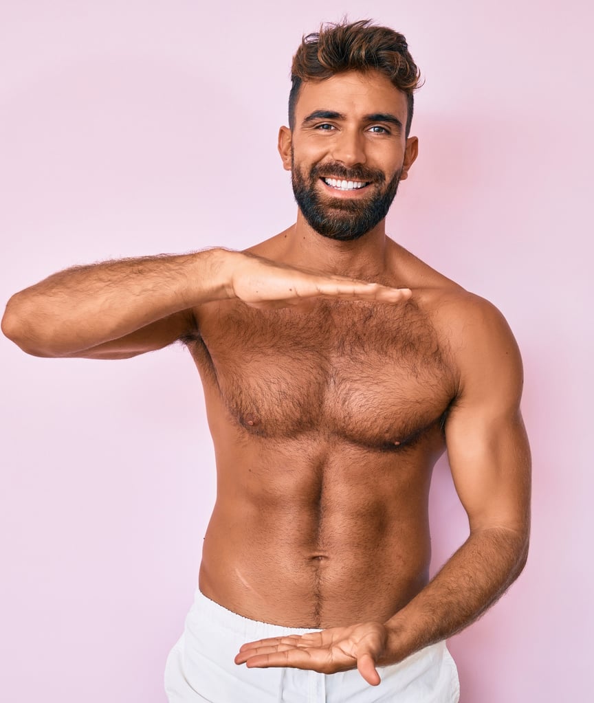 Young hispanic man standing shirtless with natural but trimmed chest hair gesturing with hands showing big and large size.