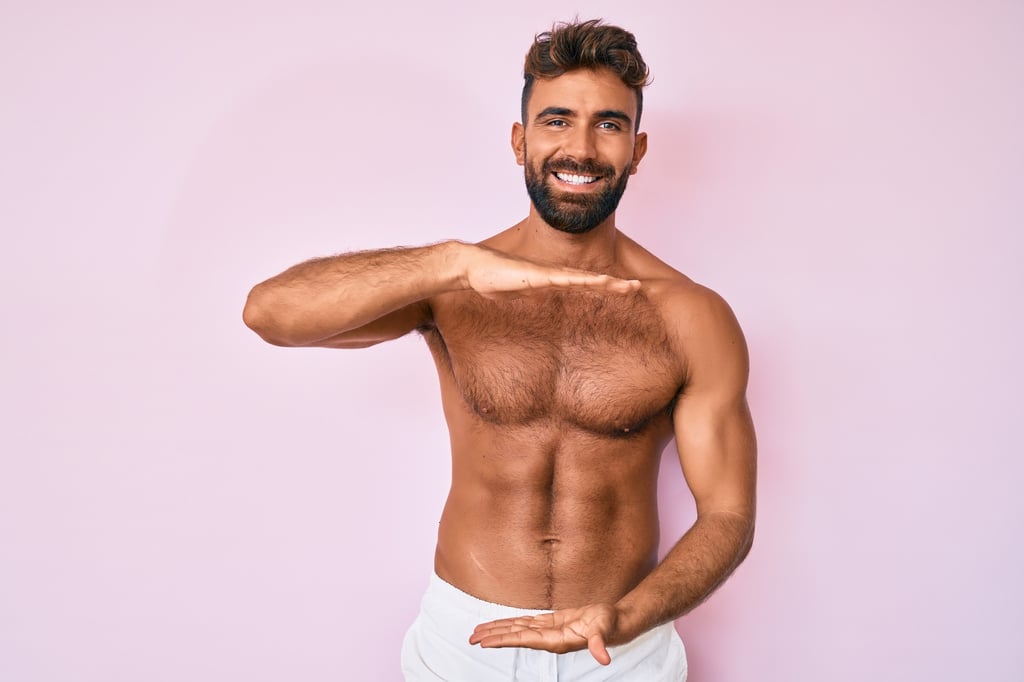 Young hispanic man standing shirtless with natural but trimmed chest hair gesturing with hands showing big and large size.