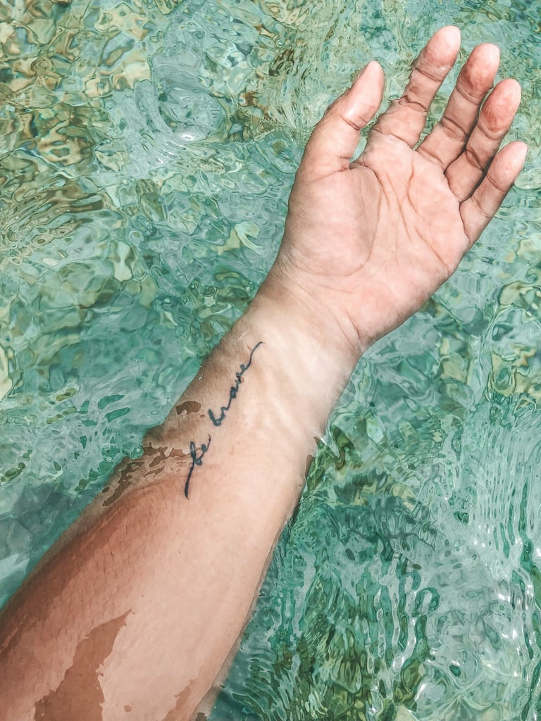 A forearm tattoo on the wrist with the phrase "be brave" written in simple script.