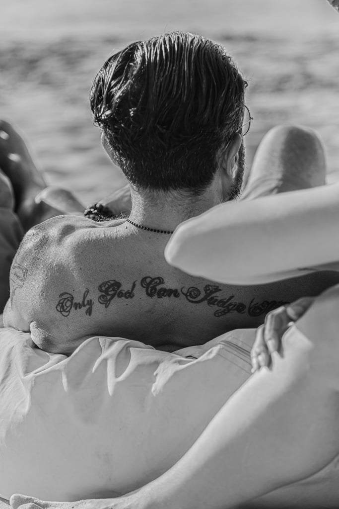 A man with damp hair has a tattoo across his upper back that reads "Only God Can Judge," with a blurred ocean in the background.