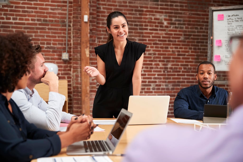 A young businesswoman standing and leading an office meeting around a table, representing leadership and the empowerment behind hustle quotes for women.