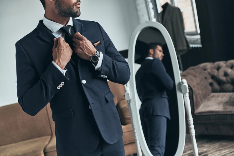 Man adjusting his suit jacket and tie while standing in front of a full-length mirror in a stylish room.