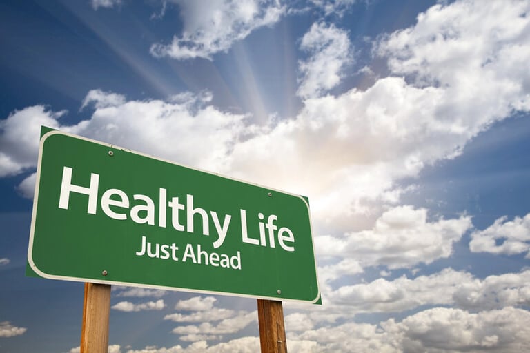 Green road sign reading “Healthy Life Just Ahead” with bright sunlight and clouds in the background