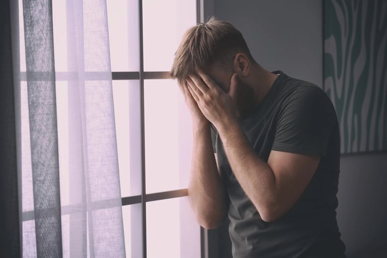 Stressed man covering face near window, symbolizing emotional distress and the importance of on-demand therapy for mental health support and crisis prevention.