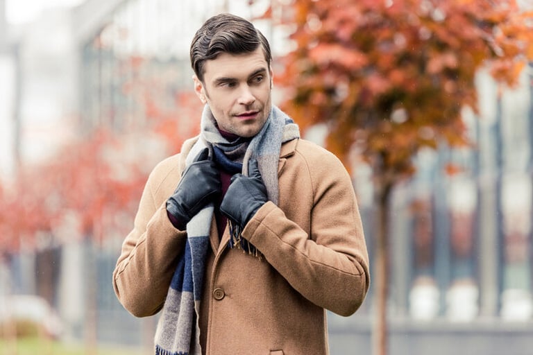A man in a camel coat and scarf standing outdoors in autumn, adjusting his gloves.