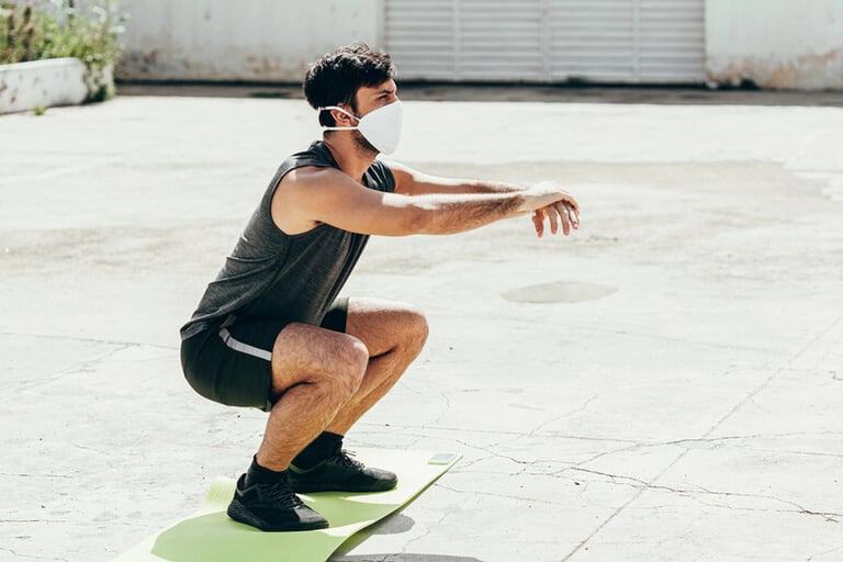 Young man wearing a face mask doing squats on an exercise mat outdoors, symbolizing healthy lifestyle habits for men to support better health and insurance outcomes.