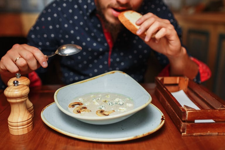 A man is eating a bowl of creamy mushroom soup and holding a piece of bread at a restaurant