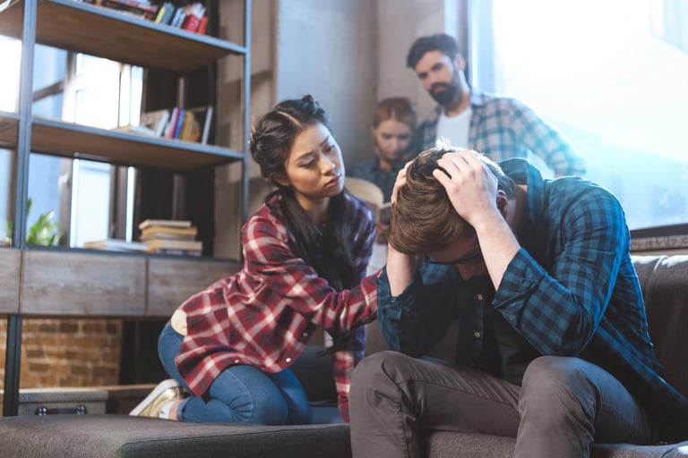 Friends who are hugging a man in distress, sitting on a couch with his head in his hands.