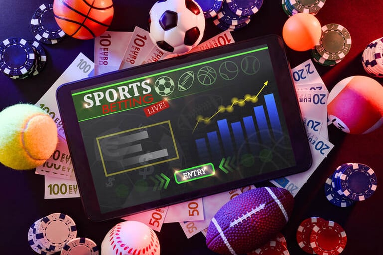 Live sports betting interface on a tablet showing real-time performance charts, surrounded by sports balls, casino chips, and cash during an in-game betting scenario.