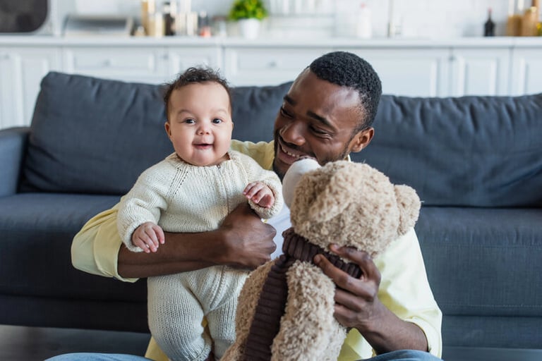 Father holding a baby and playing with a teddy bear.