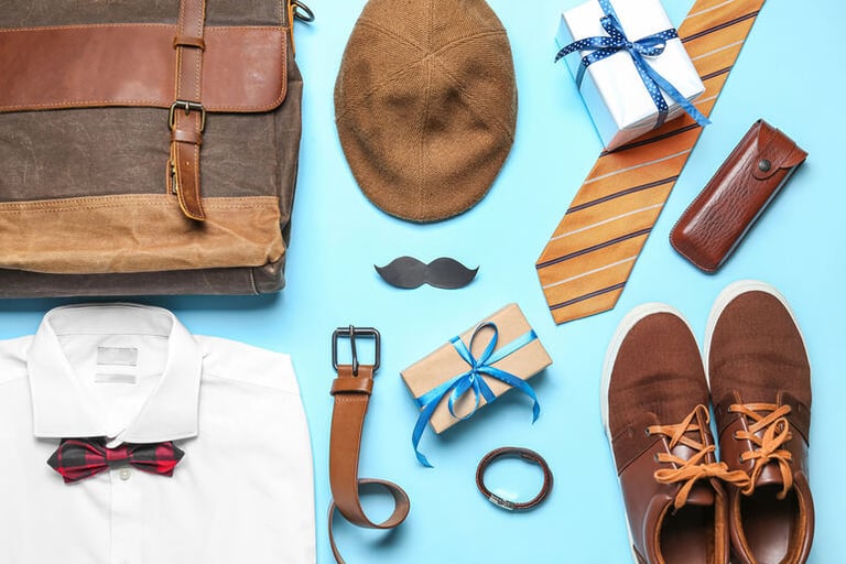 Flat lay of men’s essentials, including a leather bag, brown shoes, a shirt with bow tie, cap, tie, belt, watch, and gift boxes styled as a grown man’s Disney fan starter kit