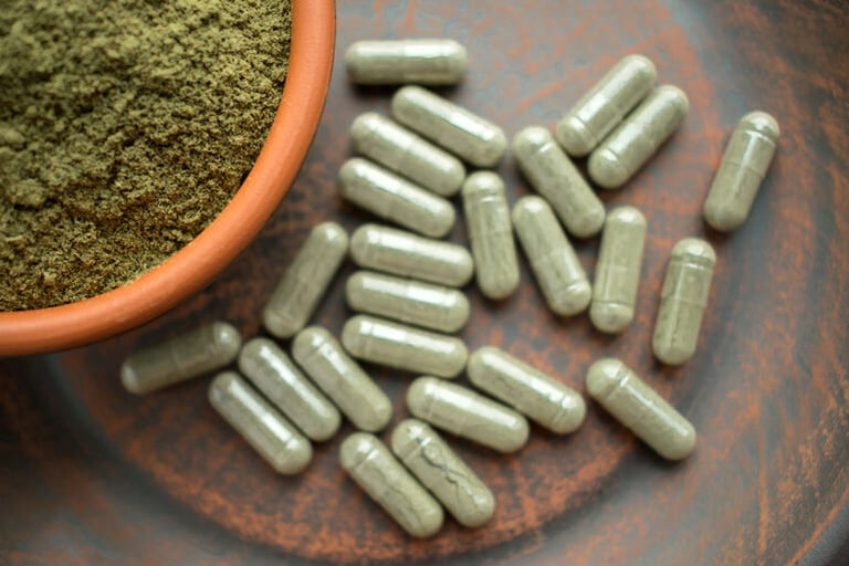 Green kratom capsules beside a bowl of finely ground kratom leaf powder.