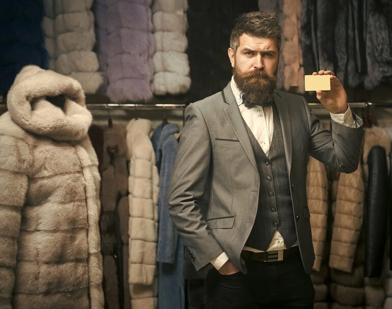 Confident, well-dressed man with a beard holding a card in a stylish clothing store, surrounded by coats, representing modern style, confidence, and personal habits