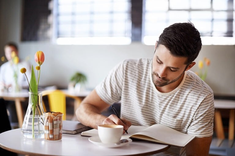 Man sitting at a table writing in a notebook beside a cup of coffee and flowers, representing simple weekly habits for staying organized and making time for reflection.