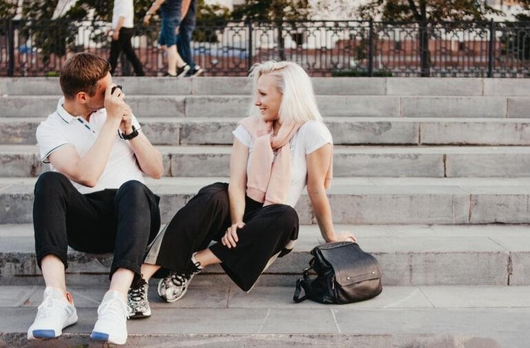 A young couple interacting on outdoor steps as the man takes a photo, illustrating a realistic connection and healthy standards.