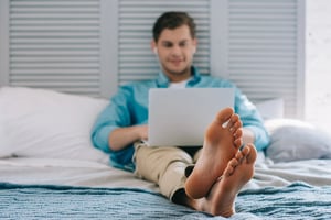 A man sitting on a bed using a laptop with his bare feet resting in the foreground.