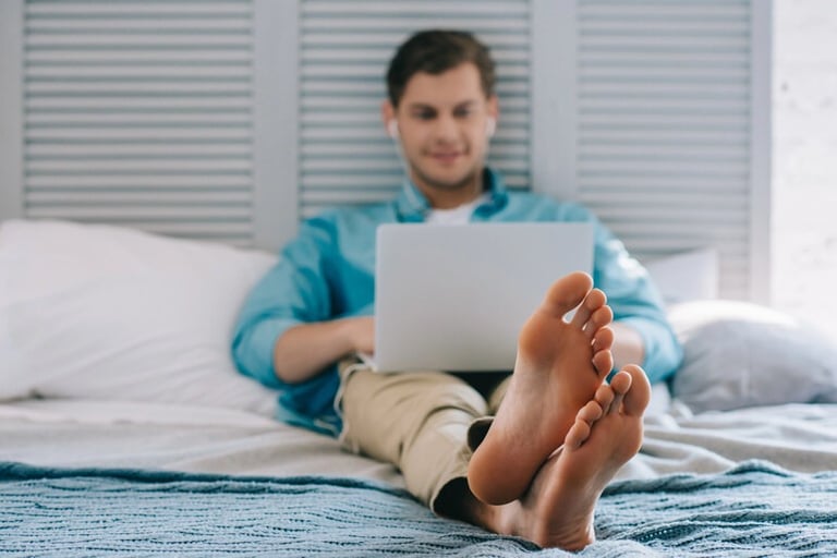 A man sitting on a bed using a laptop with his bare feet resting in the foreground.