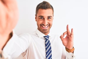 Smiling professional in business attire taking a selfie and making an OK gesture, illustrating how a confident, friendly smile helps create a positive first impression.