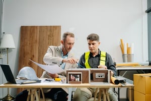 Two men collaborating on a physical architectural house model and blueprints, demonstrating hands-on career paths instead of a traditional office desk job.