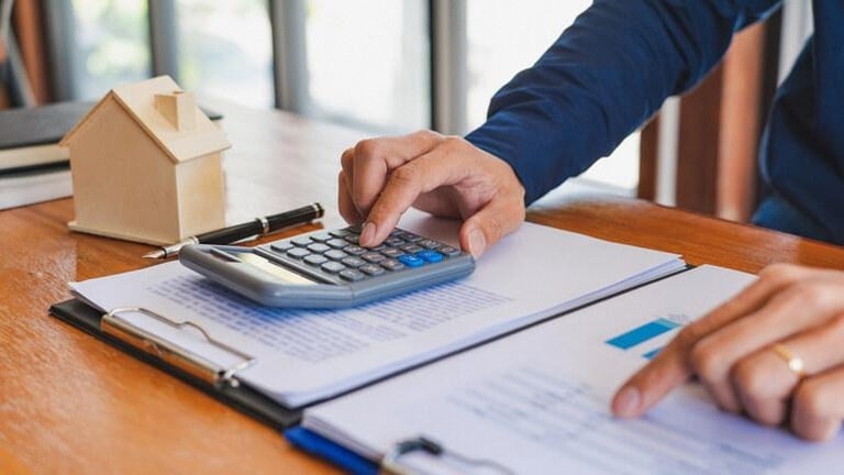Person using a calculator to estimate home loan costs with financial documents and a model house, illustrating tips for using a mortgage budgeting tool effectively.