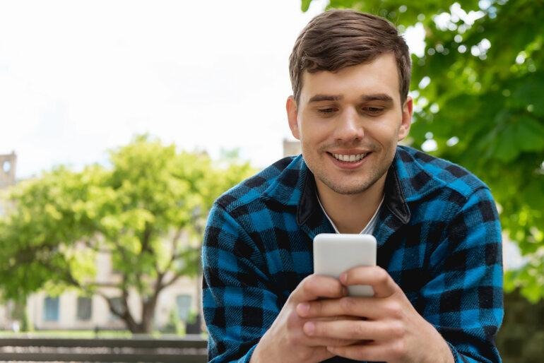 Young man smiling while texting on his smartphone outdoors, illustrating an engaging online conversation. 
