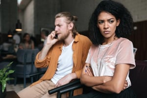 Woman sitting with crossed arms while a man talks on the phone, illustrating benching in dating and being treated as a fallback option.