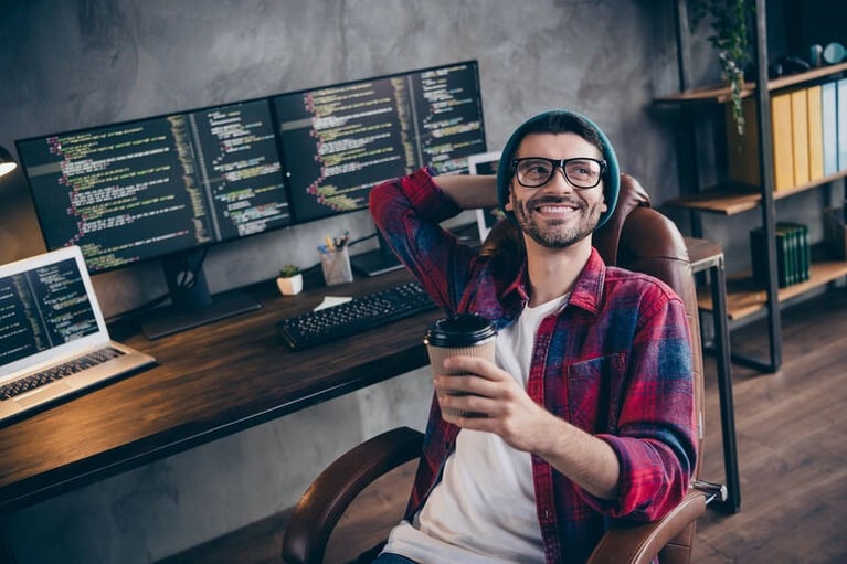 Smiling freelance web developer leaning back with coffee in front of code-filled monitors.