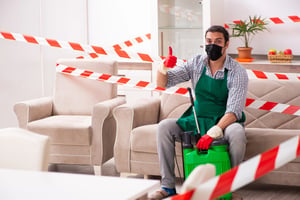 Masked pest control worker with a sprayer sitting in a living room surrounded by warning tape.
