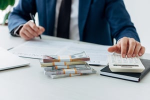 A man at a desk using a calculator and pen next to stacks of cash and financial documents.
