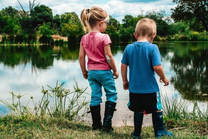 Brother and sister at the edge of a pond looking for tadpoles in the water.