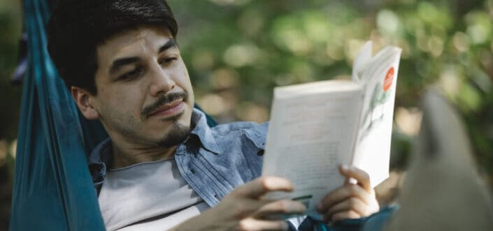 Man enjoys reading book while on a hammock.