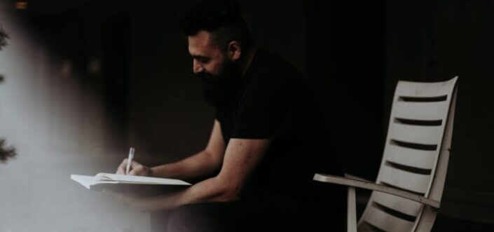 Man in black shirt writing while sitting on wooden chair.