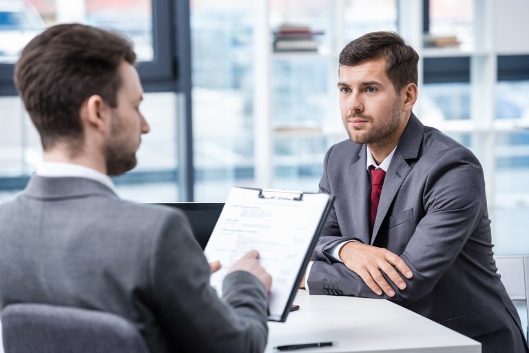 wo businessmen sitting across from each other during a job interview, with one holding a clipboard
